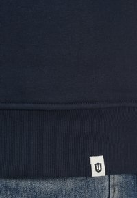 Marineblå bomulds sweatshirt med en struktureret ribbet kant og et lille hvidt label med et logo. Sættes sammen med falmede denimjeans.