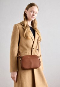 Hnědá kožená crossbody taška s hladkým povrchem, zaobleným tvarem a dvojitým předním panelem. Obsahuje dlouhý nastavovací popruh.