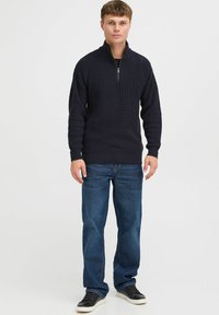 Dunkelnavy gestrickter Pullover mit einem Reißverschlusskragen, kombiniert mit blauen Jeans und schwarzen Turnschuhen. Glatte Textur und Rippmuster im gesamten Design.