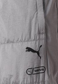 Grå letvægtsstofjakke med et sort Puma-logo og en "DRIVE" grafik, med en glat tekstur og minimalistisk design.