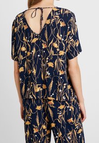 Marinblus med blommönster i gult och vitt, korta ärmar och en nyckelhålsöppning bak med en knytdetalj. Lättviktig tyg.