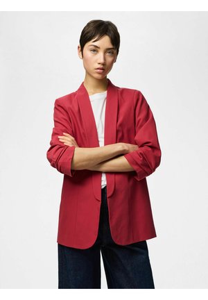 Jaunas žmogus trumpais tamsiais plaukais, vilkintis raudoną švarką virš baltos marškinėlių, susikryžiavęs rankas, stovintis priešais paprastą šviesų foną.