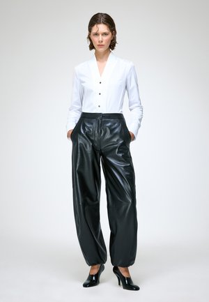 Femme debout les mains dans les poches, portant une chemise blanche boutonnée, un pantalon en cuir noir à jambes larges et des chaussures noires à talons hauts sur un fond uni.