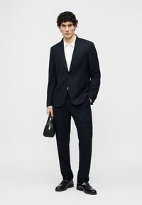 Άνδρας που φοράει μαύρο κοστούμι και λευκό πουκάμισο κρατώντας μαύρη τσάντα, όρθιος μπροστά σε απλό ανοιχτό γκρι φόντο.