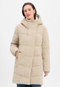 Manteau doudoune beige avec un motif texturé et matelassé, capuche et fermeture frontale diagonale. Dispose de deux poches latérales et d'une silhouette décontractée.