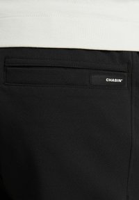 Zwarte broek met een gladde textuur, voorzien van een achterzak en een wit "CHASIN'" label. De tailleband is gedeeltelijk zichtbaar en heeft een lichte kleur.