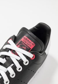 Svarta läder Adidas Stan Smith sneakers med vita snören, rosa öglor och en röd etikett med logotyp och godkännandedetaljer.