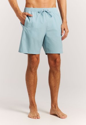 Man in lichtblauwe zwemshort met trekkoord, blootsvoets staand met één hand in zijn zak tegen een effen achtergrond.