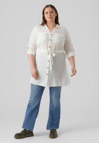 Camisa blanca de manga larga con dos bolsillos en el pecho y un cinturón, acompañada de pantalones vaqueros de campana azules y zapatos de plataforma verde oscuro.
