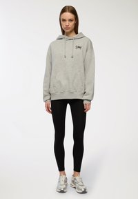 Felpa grigia con cappuccio e tasca frontale, con scritte nere; abbinata a leggings neri e scarpe da ginnastica argento su uno sfondo neutro.