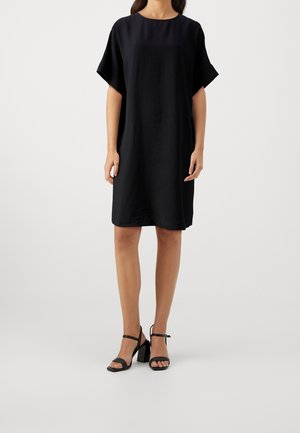 Kobieta stojąca, ubrana w luźną czarną sukienkę z krótkim rękawem do kolan oraz czarne sandały na słupku z odkrytymi palcami na jednolitym tle.