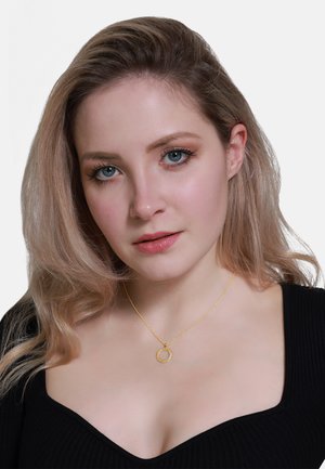 Collier pendentif en or avec un design circulaire et une texture perlée, suspendu à une chaîne délicate. Porté avec un haut en maille noir.