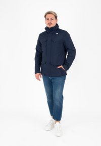 Jacke a vento blu navy con colletto alto, due tasche sul petto e chiusura con zip. Indossata con jeans blu chiaro e sneakers bianche.