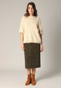 Crèmekleurige gebreide trui met wijde mouwen, gecombineerd met een groene corduroy midi-rok. Het model draagt bruine suède enkellaarsjes.