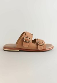 Tan ruskind slide sandal med to justerbare stropper, guldfarvede hardware-detaljer og en flad brun sål. Minimalistisk design, åben bag.