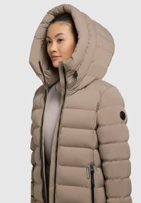 Jachetă puffer bej cu o glugă mare, ajustabilă. Caracteristici include textură matlasată, buzunare cu fermoar și o insignă rotundă pe mânecă.