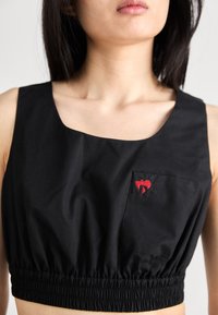Haut court noir en tissu lisse, avec un décolleté carré, une taille élastique et une poche poitrine gauche ornée d'un logo rouge brodé.
