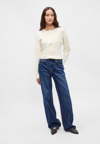 Κρεμ ριμπ κάρντιγκαν με κουμπιά στο μπροστινό μέρος, συνδυασμένο με ψηλόμεσα, φαρδιά μπλε τζην παντελόνια και μαύρες μυτερές γόβες με ασημί λεπτομέρειες.