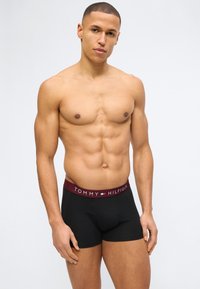 Μυώδης άνδρας που φορά μαύρα boxer briefs Tommy Hilfiger, στέκεται μπροστά σε απλό φωτεινό φόντο, στραμμένος προς τα εμπρός.