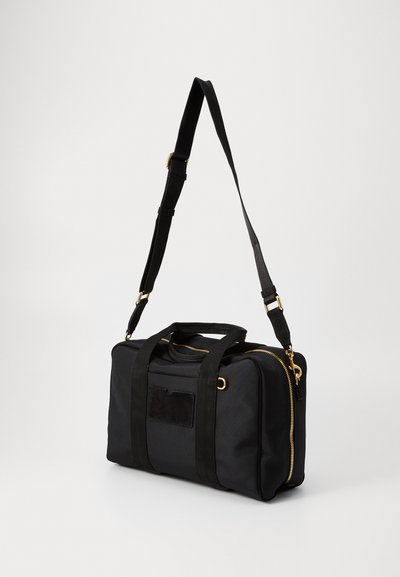 Sac de sport en tissu noir avec fermeture éclair dorée, deux poignées supérieures, une bandoulière réglable et un accent texturé noir à l'avant.
