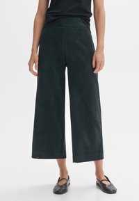 Dark green wide-leg trousers made of a textured material, featuring a high waist and cropped hem, paired with flat black shoes.