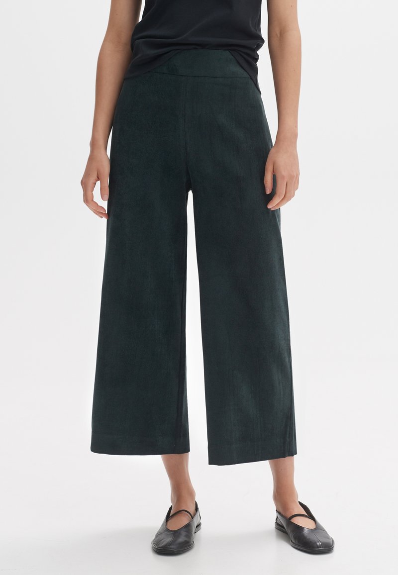 Dark green wide-leg trousers made of a textured material, featuring a high waist and cropped hem, paired with flat black shoes.