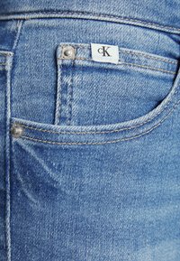 Ljusblå denimjeans med en synlig ficka, silverdetaljer och en vit etikett med "cK"-logotypen. Sliten textur och sömnadsdetaljer.