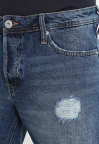 Modré džínové kalhoty s vybledlým záplatou a opotřebovanými detaily v okolí stehna, se stříbrným knoflíkem a tradičním pětikapsovým designem.