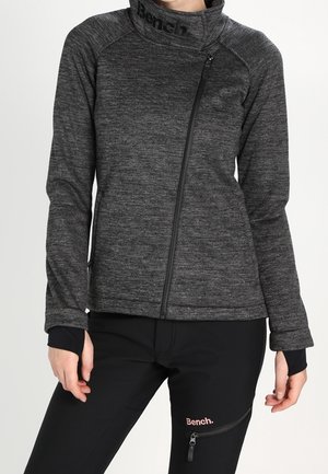 Persoon draagt een donkergrijs jack met rits en hoge kraag en zwarte broek met ritszak en Bench-logo op beide kledingstukken.