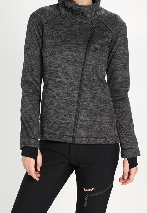 Personne portant une veste zippée gris foncé à col montant et un pantalon noir avec une poche zippée et le logo Bench sur les deux vêtements.