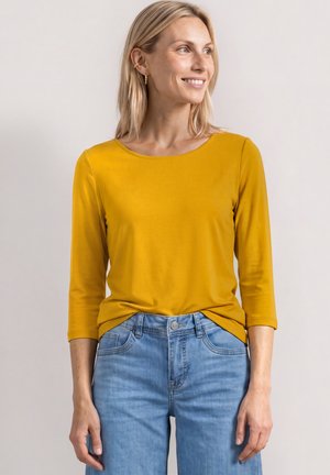 Kvinde med blondt hår, der smiler og kigger sidelæns iført en sennepsgul langærmet top og lyseblå jeans.