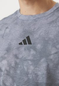 Pilkas sportinis marškinėliai su tekstūruotu raštu, apvaliu iškirpimu ir juodu Adidas logotipu priekyje, demonstruojantis laisvalaikio dizainą.
