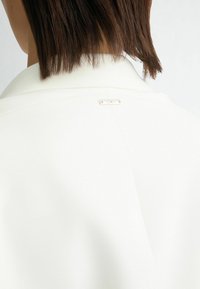 Witte blazer met een gladde textuur, voorzien van een subtiele kraag en een klein gouden hardware-detail nabij de achterkant van de halslijn.