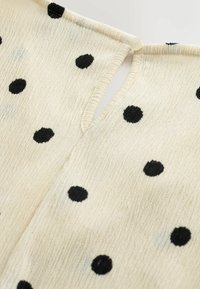 Tissu de couleur écrue à pois noirs, avec une petite boutonnière ronde au sommet d'un décolleté en forme de goutte d'eau.