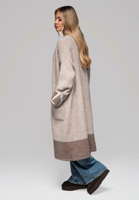 Maglione lungo e ampio in grigio chiaro con orlo crema e marrone. Presenta accenti decorativi sulle maniche e sulle tasche laterali. Tessuto dalla texture morbida.