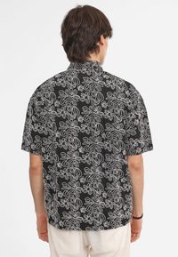 Camicia nera a maniche corte con un motivo floreale bianco, orlo dritto e colletto con bottoni. Tessuto morbido con una texture liscia.