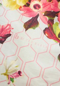 Ziedu audums ar lieliem rozā, dzelteniem un violetiem ziediem pār rozā sešstūra rakstu un vieglu "by Ted Baker" tekstu.