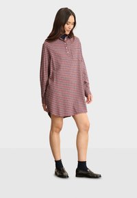 Robe chemise à carreaux rouges et bleus avec des manches longues, un plastron à boutons et un ourlet arrondi. Associée à des chaussettes sombres et des mocassins.