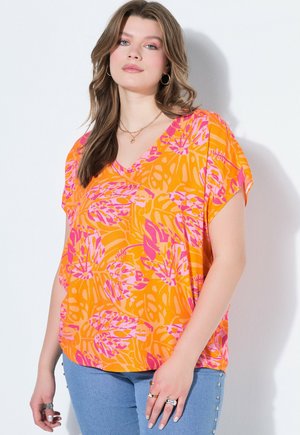 Kobieta w pomarańczowo-różowej kwiecistej bluzce z dekoltem w serek i krótkimi rękawami oraz w niebieskich dżinsach z perełkowymi ozdobami, stojąca przy białej ścianie.