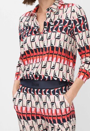 Nainen pukeutuneena yhteensopivaan, punaisen, pinkin, mustan ja valkoisen geometrisen kuvion napitettuun paitaan ja housuihin, kädet taskuissa.