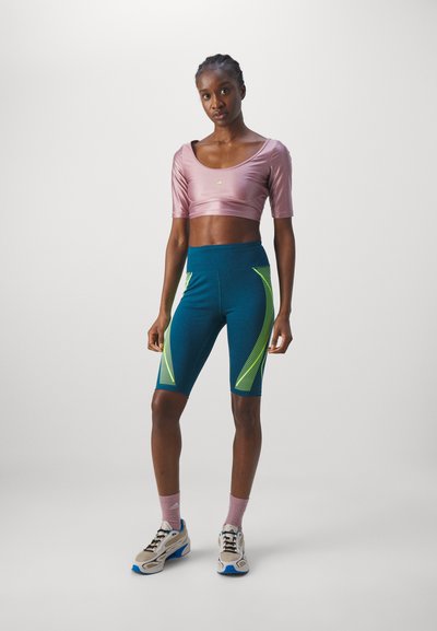 En atletisk kvinne står og har på seg en skinnd pink crop top, blå sykkelshorts med grønne mønstre, rosa sokker og hvite joggesko.