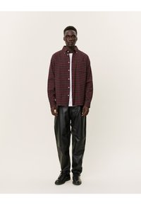 Camicia a quadri rossa e verde, con maniche lunghe, abbinata a pantaloni larghi in pelle nera e sneaker nere. Design ad alto contrasto con una vestibilità rilassata.