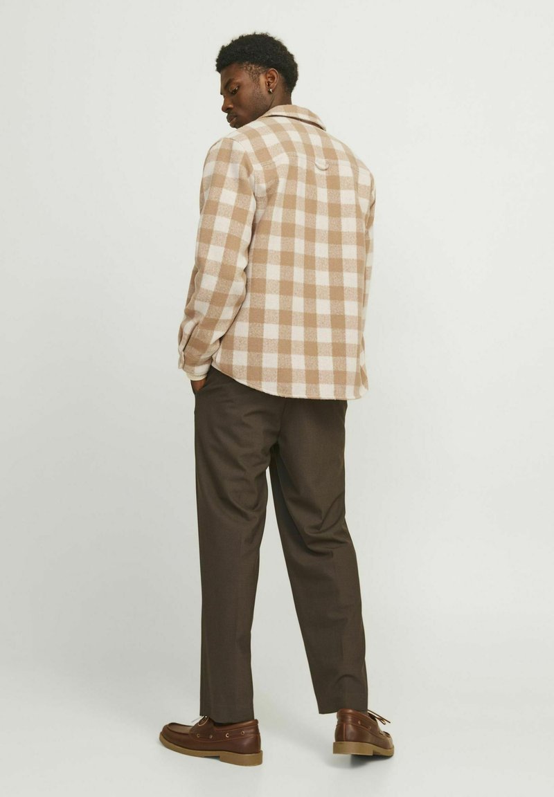 Brown and cream checkered flannel shirt, button-up style with a rounded hem, paired with dark brown trousers and brown lace-up shoes.