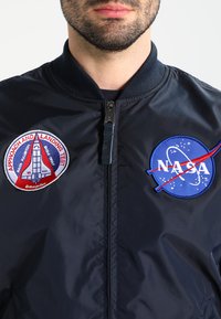 Mørkeblå bomberjakke med ribbet krave, prydet med NASA og rumfærge-mission patches i rød, hvid og blå broderi. Glat, skinnende materiale.