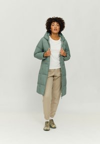 Long, lightweight puffer coat in muted green, featuring a hood, zip closure, and quilted design. Paired with cream joggers and multi-colored shoes.