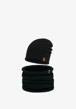 Mosolygó nő, fekete kötött sapkát és hozzá illő vastag fekete kötött nyakmelegítőt visel bézs kabát fölött.