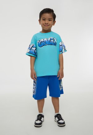 T-shirt grafico turchese con accenti blu, caratterizzato dal testo "CALI." Abbinato a pantaloni corti blu solidi con un design coordinato, indossati con scarpe da ginnastica nere.