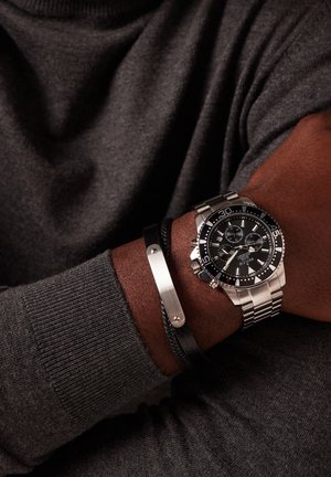 Edelstahl-Uhr mit schwarzem Ziffernblatt, silbernen Akzenten und Chronographenfunktionen. Kombiniert mit einem schwarzen geflochtenen Armband mit Metallverschluss.