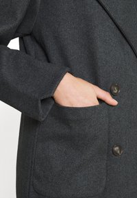 Mörkgrå herringbone-kappa med en enkel knappstängning och fickor; tyget verkar texturerat och mjukt.