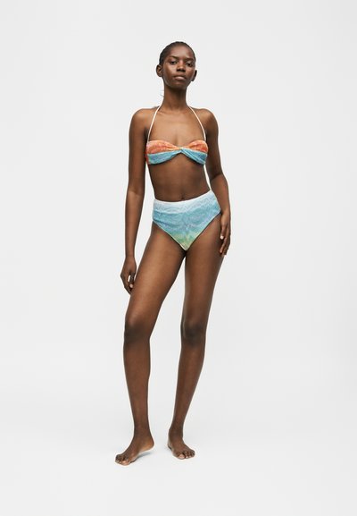 Femme portant un bikini bandeau à col licou avec un haut orange et bleu et un bas taille haute bleu et vert, se tenant sur un fond blanc.